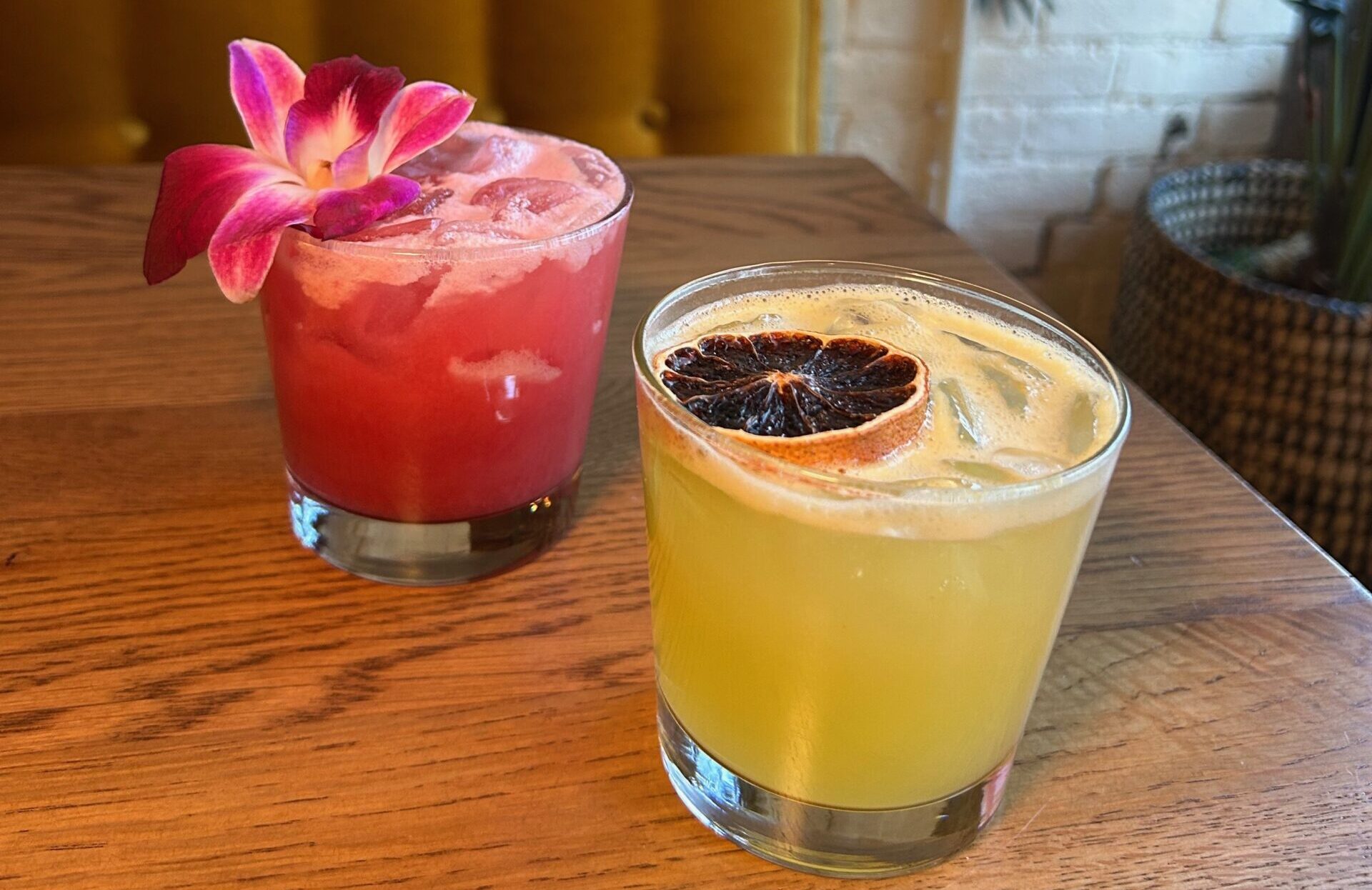 two cocktails on a table. one is pink with a flower and the other is yellow with an orange slice